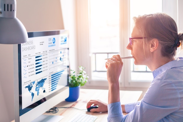  A woman in glasses is seen in profile from the waist up, sitting at a desk and looking at a large computer monitor that shows several charts under the label "Business Dashboard." The charts include a map, a few bar graphs, and a small pie chart, all in various shades of blue. The woman in glasses uses one hand to manipulate a corded computer mouse and the other hand to holds a pen to her mouth in thought.