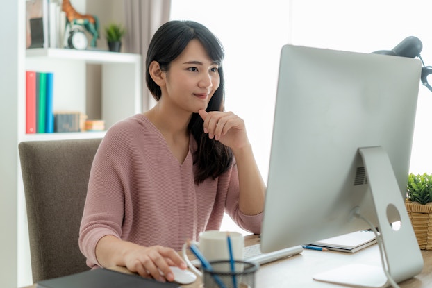 A young woman with long dark hair sits at a desk and looks at a large computer monitor with a slight smile. She has one hand on a wireless mouse and the other held to her chin.
