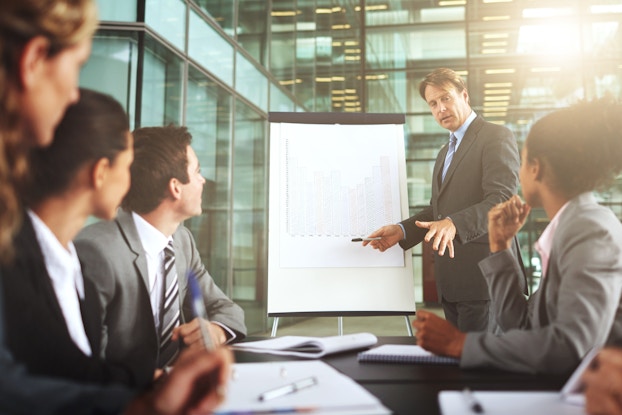  A man in a suit stands mid-speech and pen in hand next to a large easel on which is mounted a bar chart. In the foreground, five people in business casual clothes sit at a table with pads of paper and pens in front of them. Their heads are turned away from the camera and toward the man speaking.