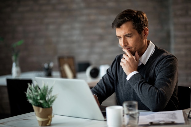 A man sits at a desk in front of an open laptop with one hand near his chin in thought. The man has dark hair and a stubbled face. He wears a dark gray sweater over a white collared shirt. In the background, out of focus, is another desk and a gray exposed brick wall.