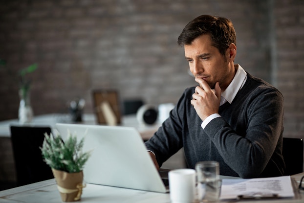  A man sits at a desk in front of an open laptop with one hand near his chin in thought. The man has dark hair and a stubbled face. He wears a dark gray sweater over a white collared shirt. In the background, out of focus, is another desk and a gray exposed brick wall.