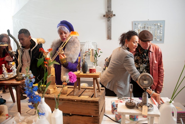 Several customers gather around a pop-up store to inspect various items for sale. The shop's owner, a woman, talks to one customer about an item he is interested in. She is gesturing to an item on display.