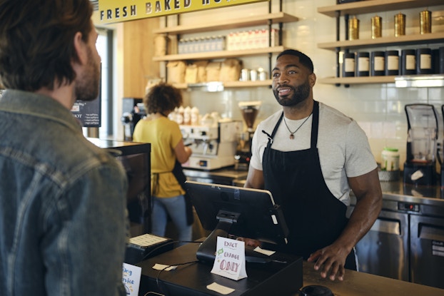  A man in a black apron stands behind a cash register in a coffee shop. A customer in a denim jacket stands across from him, facing away from the viewer. The cash register sits on a wooden counter and is made up of a large cash drawer and a mounted digital tablet. Behind the tablet, facing the customer, is a small sign reading "Exact Change Only." Behind the cashier, another employee, out of focus, works at a chrome coffee machine.