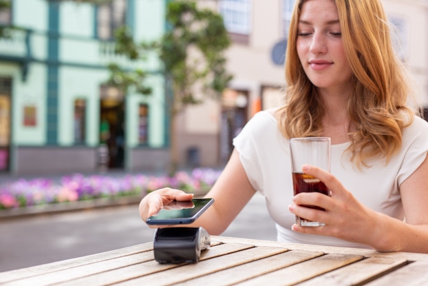A young woman holds her smartphone above a handheld point-of-sale unit. She is sitting at a wooden cafe or restaurant table outside and holding a drink in her free hand.