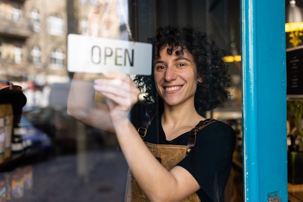 A smiling person with dark, curly hair stands behind a window and hangs a small sign that bears the word "OPEN." The room behind the person is obscured by a reflection in the window, which shows a building across the street.