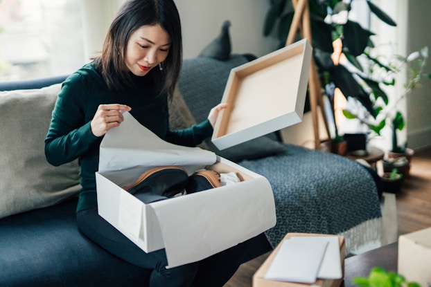 A woman sits on a couch and opens a box. In one hand, she holds the box's lid; with the other hand, she pulls back tissue paper to reveal a pair of black boots with rubber soles.