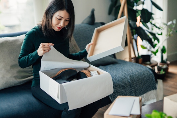  A woman sits on a couch and opens a box. In one hand, she holds the box's lid; with the other hand, she pulls back tissue paper to reveal a pair of black boots with rubber soles.