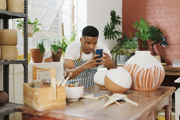 A young man taking pictures of his finished vases with a smartphone in a pottery studio.
