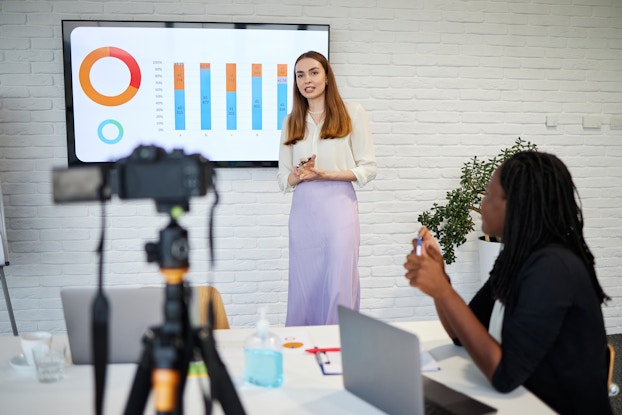  A woman stands in front of a whitewashed brick wall on which is mounted a flat screen TV. The TV shows two graphs -- an orange-and-red circle graph and a blue-and-orange bar graph. In the foreground, another woman sits at a table and watches the presentation. A camera on a tripod is pointed at the presenter.