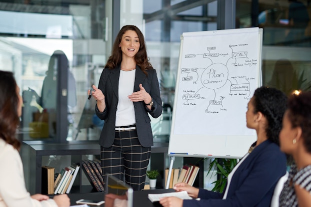  A woman in a dark blue blazer and black plaid pants stands in front of a glass wall and next to a large pad of paper on an easel. The paper shows a chart with several labels branching off of a central circle labeled "Plan." In the foreground, four women of various races watch the first woman talk.