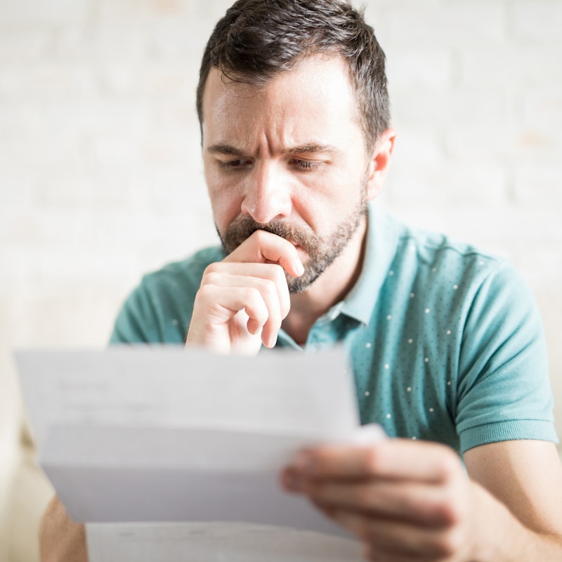 A bearded man sits on a cream-colored sofa and looks at a creased piece of paper with a worried expression.