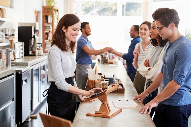 A woman behind the counter at a coffee shop smiles as she rings up a customer. Multiple customers wait in line for their orders.