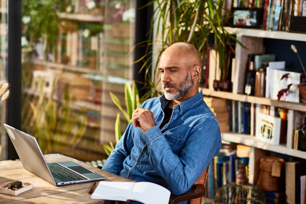  A man sits at a table in front of an open laptop with a thoughtful look on his face. The man is wearing a chambray shirt and has a shaved head and a salt-and-pepper beard. An open book sits next to the laptop on the table, and shelves of books stand behind the man.