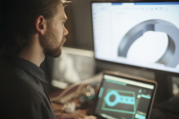 A man, facing away from the camera at an angle, looks at two computer screens. The first screen is on a laptop and shows a black-and-teal-blue design of a key-shaped machine component made up of an open circle attached to a rectangular piece. The second screen sits behind the laptop and is a much larger monitor. It shows a zoomed-in 3D look at the circular part of the component.