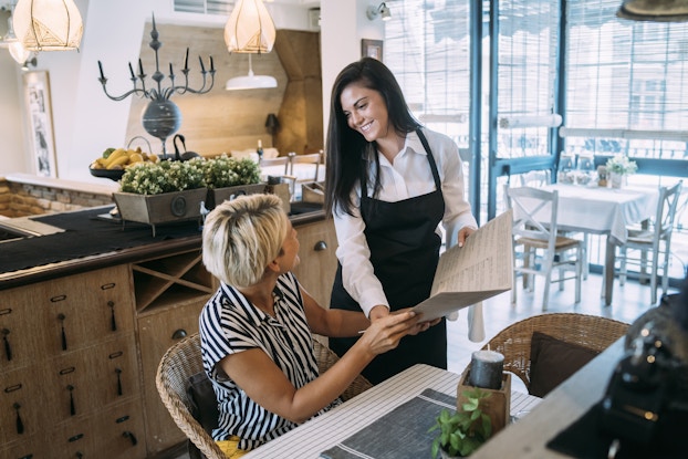A diner and a server in an upscale restaurant. The server, wearing a black apron, holds a menu open and smiles at the customer. The customer, a woman with short, light hair, is sitting at a table in a wicker chair and looking up at the server.