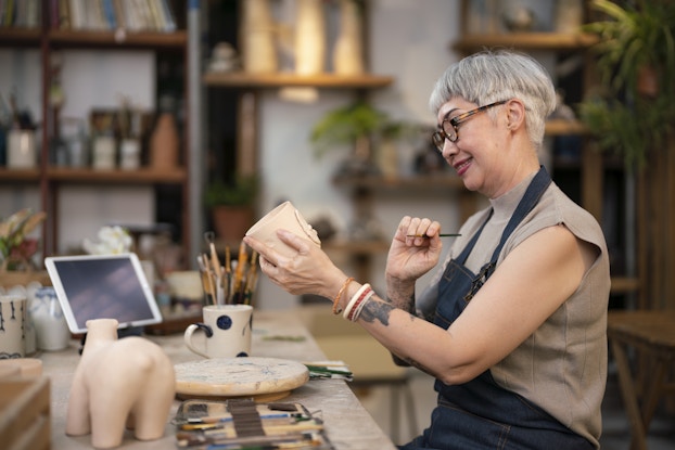 An older woman sits at a worktable in a ceramics workshop and paints details on a ceramic mug. She holds the mug in one hand and looks down at it with a smile. She holds a thin paintbrush in the other hand. The woman has short light gray hair and wears a pale beige T-shirt, a dark blue apron, and round tortoiseshell glasses.