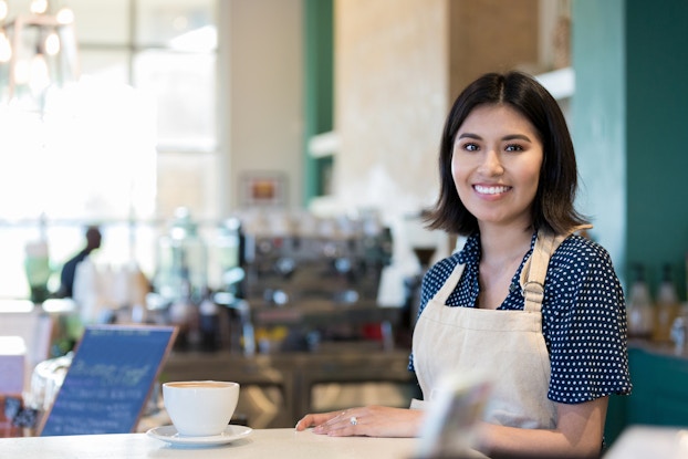 A young woman wearing a khaki apron stands behind a counter at a coffee shop. A large white cup of coffee sits on a saucer on the counter in front of her. In the background, out of focus, are a small chalkboard with chalk writing on it and various implements used for brewing and serving coffee.