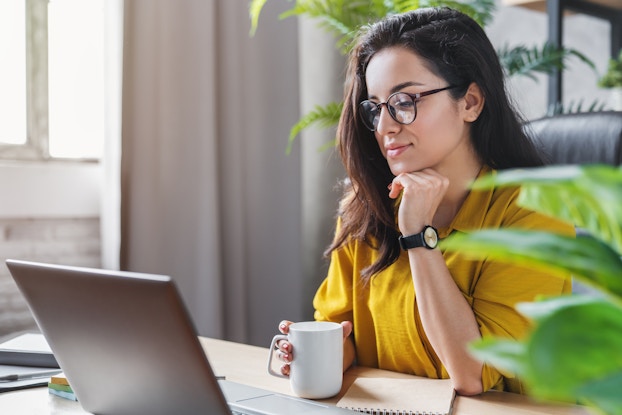 A woman in a yellow shirt sits at a desk and looks at the screen of an open laptop. One of her hands is curled under her chin and the other is holding a white mug.