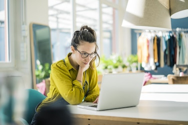 A woman in a yellow-green button-up shirt sits at a table and looks at the screen of a laptop. The woman has her long dark hair pulled up into a ponytail and she wears glasses. In the background is a rack of clothing and a large mirror.