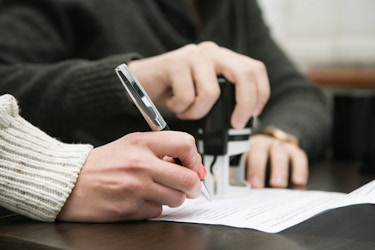  Close-up on two sets of hands. One person's hand signs a contract while the other person readies a stamp for the form. 
