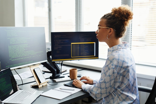 A woman sits at a desk in an office and faces two large computer monitors. The monitors both show black screens covered with lines of coding text. The woman wears glasses and a white-and-blue patterned shirt. She has both hands on a computer keyboard. Next to the keyboard on the desk are an open notebook, an open laptop, and a smartphone sitting on a stand.
