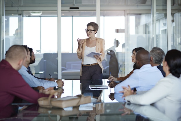  A young woman with short hair stands at the front of a conference room and speaks to her colleagues, who are seated around a mirror-topped conference table.