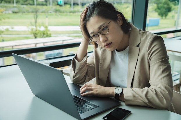 A tired-looking young woman sits in front of an open laptop with her head leaning on one hand. She's sitting beside a floor-to-ceiling window, through which can be seen a grassy area bisected with a white pathway. The woman wears glasses and a tan jacket over a white blouse.