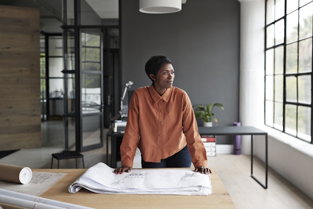 Black woman entrepreneur looking over blueprints and gazing out her office window.