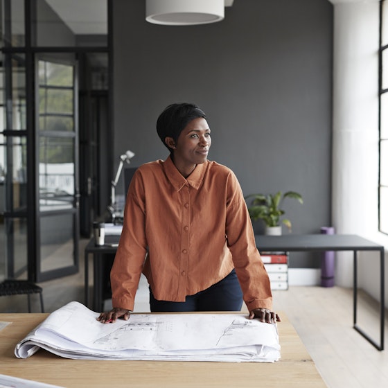 Black woman entrepreneur looking over blueprints and gazing out her office window.