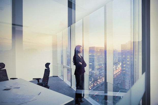  Woman dressed in a business suit inside an office staring out of the floor-to-ceiling windows.