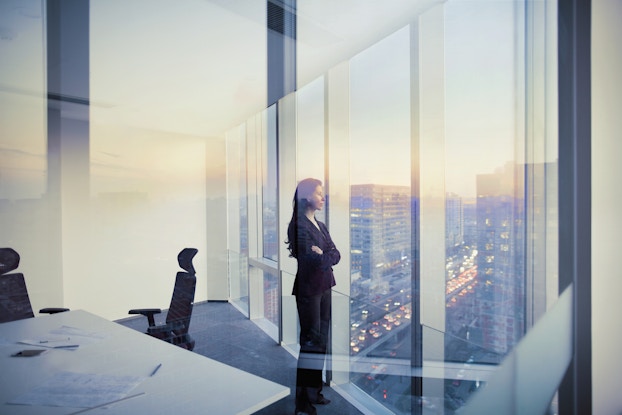 Woman dressed in a business suit inside an office staring out of the floor-to-ceiling windows.
