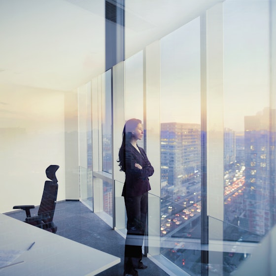 Woman dressed in a business suit inside an office staring out of the floor-to-ceiling windows.