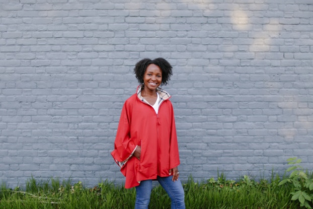  Woman smiles as she stands in front of a white brick wall and grass, wearing a dark orange rain jacket.