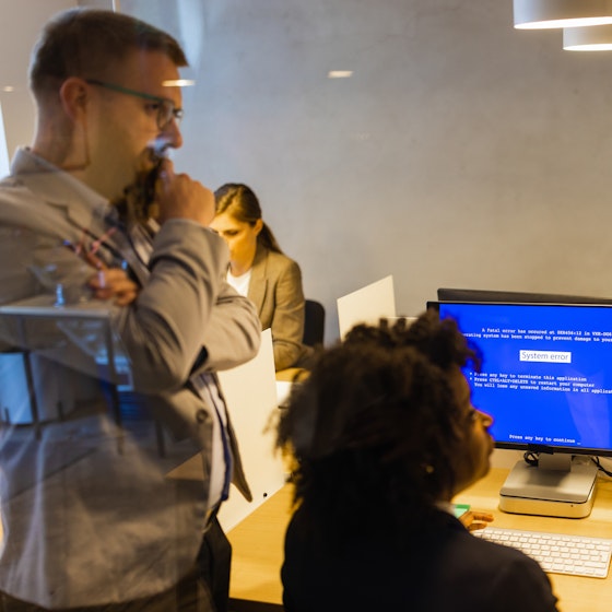  A group of people seen through a window, with reflections of lights laying over the scene. Three worried colleagues cluster around a computer showing a blue error screen. In the background, two other people work at their own computers. 