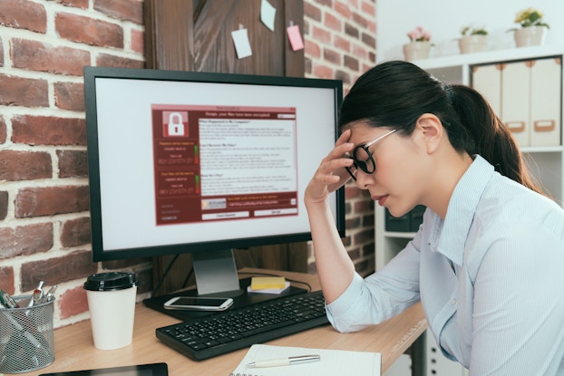  A woman sits at a desk with one hand to her forehead and her eyes closed. She looks defeated. The computer monitor on her desk displays a red and white window filled with text. An icon of a closed padlock suggests that the computer is locked.