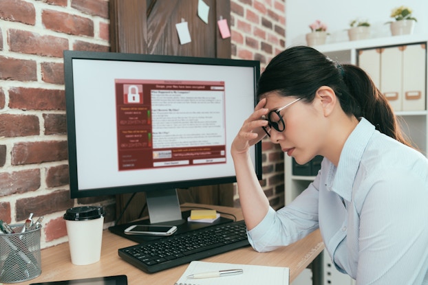 A woman sits at a desk with one hand to her forehead and her eyes closed. She looks defeated. The computer monitor on her desk displays a red and white window filled with text. An icon of a closed padlock suggests that the computer is locked.