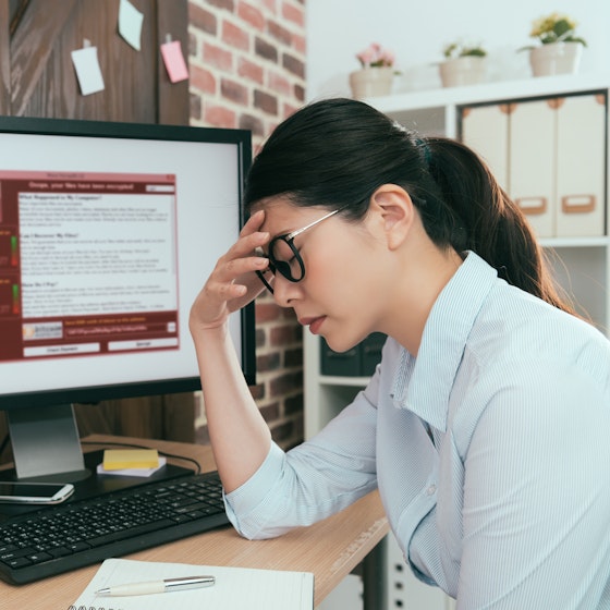  A woman sits at a desk with one hand to her forehead and her eyes closed. She looks defeated. The computer monitor on her desk displays a red and white window filled with text. An icon of a closed padlock suggests that the computer is locked. 