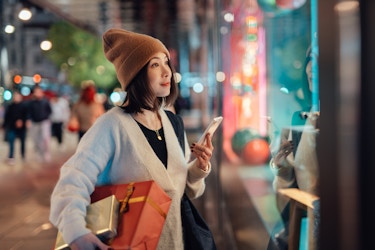  A young woman, seen in angled profile, stands outside of a shop and gazes in awe at something in the display window. The woman wears a light gray coat and brown hat, and she carries wrapped presents under one arm. In her free hand, she holds a smartphone. The items in the display window can't be seen from the viewer's angle, but the window reflects lights from the street in the background. 