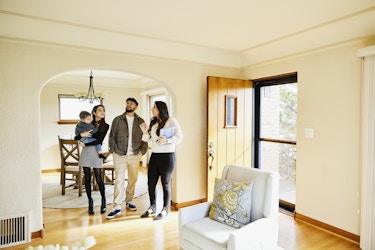  A realtor leads a family of three through a house. The group stands under the archway leading from a dining room into a living room. A table and chairs and a chandelier can be seen behind the group in the dining room, and a front door stands open in the living room. The walls of the rooms are painted cream and the floor is laminated light-colored wood. The realtor is a young woman with long, dark hair; she is holding a folder and speaking to the family. The family is made up of a young bearded man, a young dark-haired woman, and a boy around age 2 or 3. The woman carries the boy, and all three family members look around the room. 