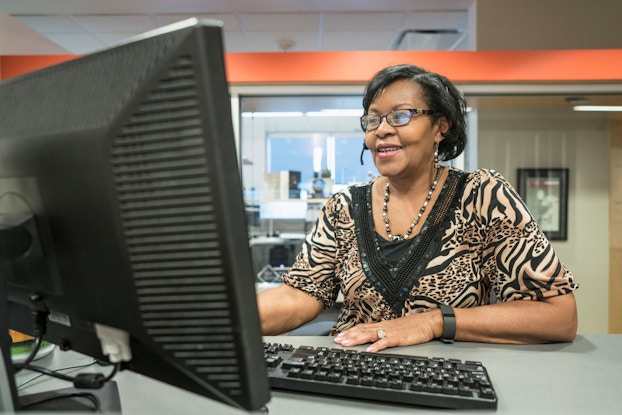 A woman receptionist sitting at her computer wears a phone headset and talks to a customer while checking her computer.