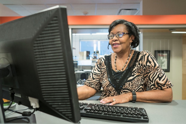  A woman receptionist sitting at her computer wears a phone headset and talks to a customer while checking her computer.