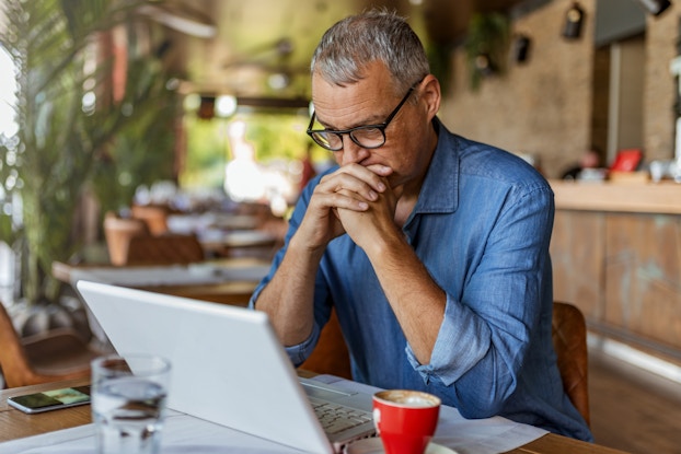  A man sits in an empty cafe in front of an open laptop. He has gray hair and is wearing glasses and a chambray shirt. His hands are folded and held to his mouth in concern.