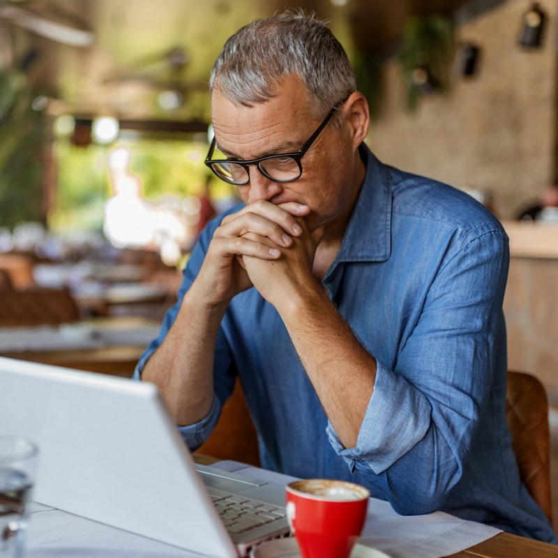 A man sits in an empty cafe in front of an open laptop. He has gray hair and is wearing glasses and a chambray shirt. His hands are folded and held to his mouth in concern. 