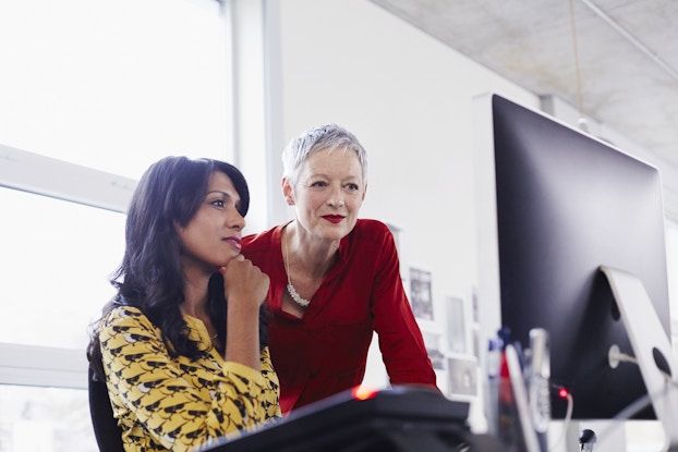 Two women, one younger and one older, look at a computer monitor in an office. The younger woman is sitting down and has one hand to her chin in deep thought. She has long dark hair and wears a yellow-and-black shirt. The older woman stands, leaning over the younger woman. She has short silver hair and wears a red shirt and red lipstick.