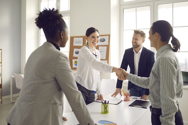  Four people wearing business casual clothes stand around a table in a white-walled room. Two of the people, both women standing on opposite sides of the table, shake hands. The other two people, a smiling man with blond hair and a beard and a dark-haired woman facing away from the camera, look on. The table holds two cups filled with pens and a couple of print-outs of pie charts. A bulletin board on the wall behind the group is covered with more print-outs of pie charts and papers covered with text.