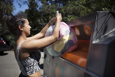  A woman seen in profile lifts a semi-transparent trash bag filled with clothes into a large metal recycling container. The woman has dark hair pulled back into a bun and she's wearing a black-and-white crop top. The recycling container is taller than the woman and about the same width as a Dumpster. It's colored dark gray, except for the wide disposal opening, which is orange. 