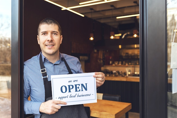  A man wearing an apron stands in the entry way of his business. In the background, the store is outfitted with wood-top tables and shelves of dark bottles. The man holds a sign that says "we are OPEN" and "supporter small businesses." "OPEN" is written in bold blue letters, with the other words arranged above and below in blue cursive.