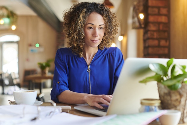 A curly-haired woman in a blue zip-up shirt is typing on a laptop at a table in a large cafe. The cafe behind her is empty, except for two people, one standing and one sitting, in the far background. On the table next to the woman's laptop is a three-ring binder filled with papers and a teacup on a saucer. A small decorative potted plant sits behind the laptop.