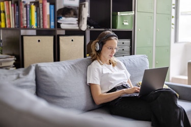  A young woman is seated on a couch in her apartment. She is wearing headphones and is typing on a laptop computer placed on her lap. 