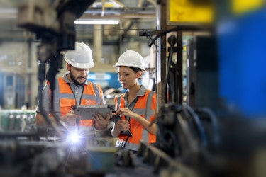 Two manufacturing workers look at the screen of an electronic tablet. The worker on the left is a bearded man, and the worker on the right is a woman with dark hair. Both workers wear gray polo shirts, white hard hats, and orange high-visibility vests. In the foreground, out of focus, is a large piece of machinery with a mechanical arm emitting sparks.
